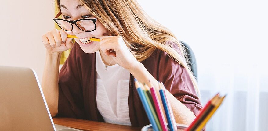 Mädchen mit Brille beißt auf einen Stift und sitzt vor einem Laptop